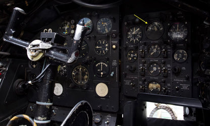 Close-up of an aircraft instrument dashboard with multiple round gauges, dials, and controls, including a yellow arrow pointing to one gauge on the right panel.