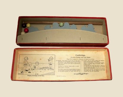Vintage wooden cuebridge game set in its original red box, with the open lid displaying the wooden bridge, blue sky cutout, and colored cue balls, and the bottom panel showing the printed instruction booklet insert.